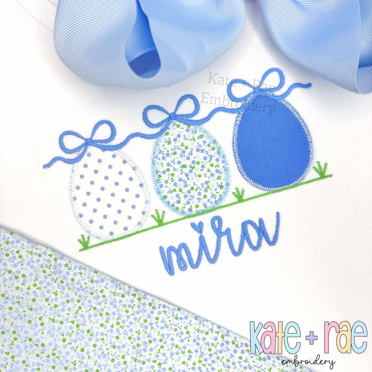 Girl's Easter Egg Trio Zigzag Applique Embroidery Design: Three blue polka dot and floral applique Easter eggs with bows above the name Mira in blue on a white shirt