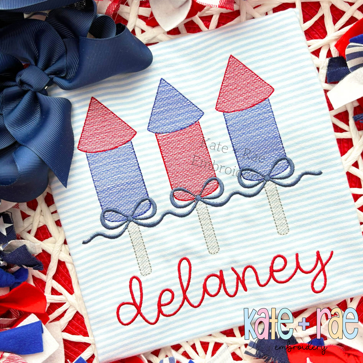 Girl's Firework Trio with Bows Sketch Stitch Embroidery Design: Three red and blue sketch firework rockets with navy bows above the name delaney in red script on a striped shirt