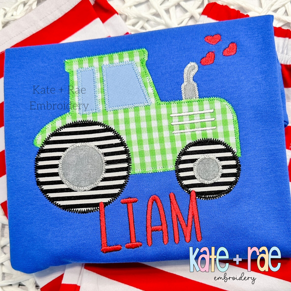 Tractor with Hearts Zigzag Stitch Applique Embroidery Design: Green gingham tractor applique with striped wheels and hearts above the name LIAM in red on a blue shirt