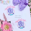 Floral Triple Flowy Bow Crest Embroidery Design: Two white baby items with pink triple bow frames and floral clusters around purple monograms