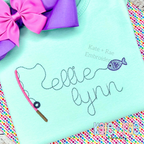 Gone Fishing Bean Stitch Embroidery Font: The name ellie lynn in gray script with a pink fishing pole and purple fish on a mint shirt