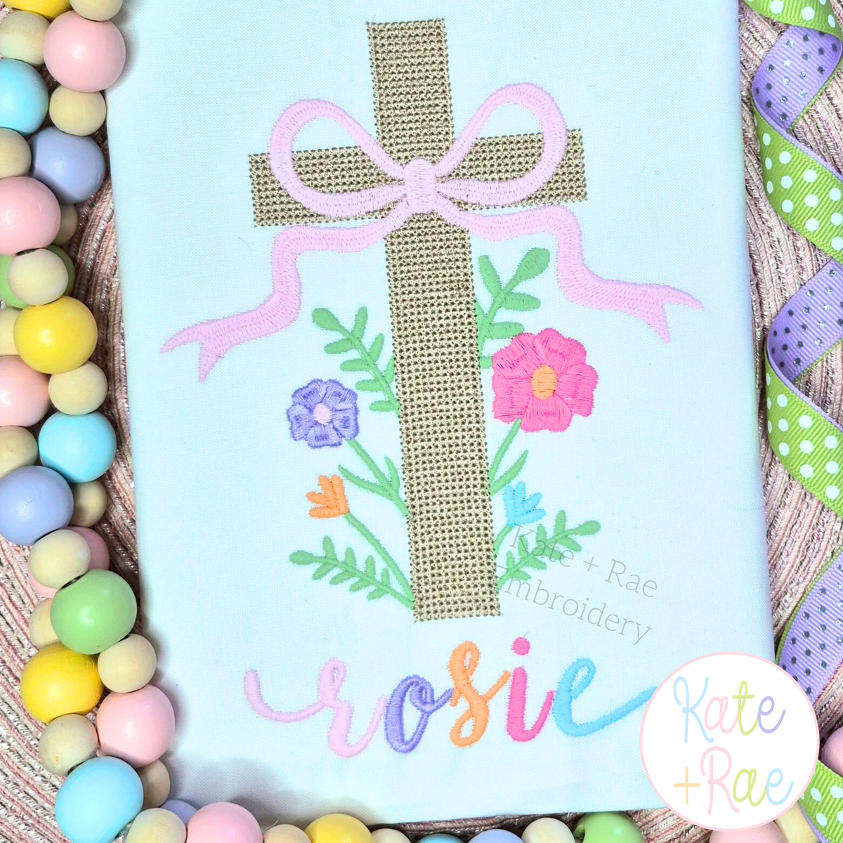Cross Stitch Floral Cross Embroidery Design: Tan cross stitch floral cross with a lavender bow above the name rosie in pastel letters on a light blue shirt
