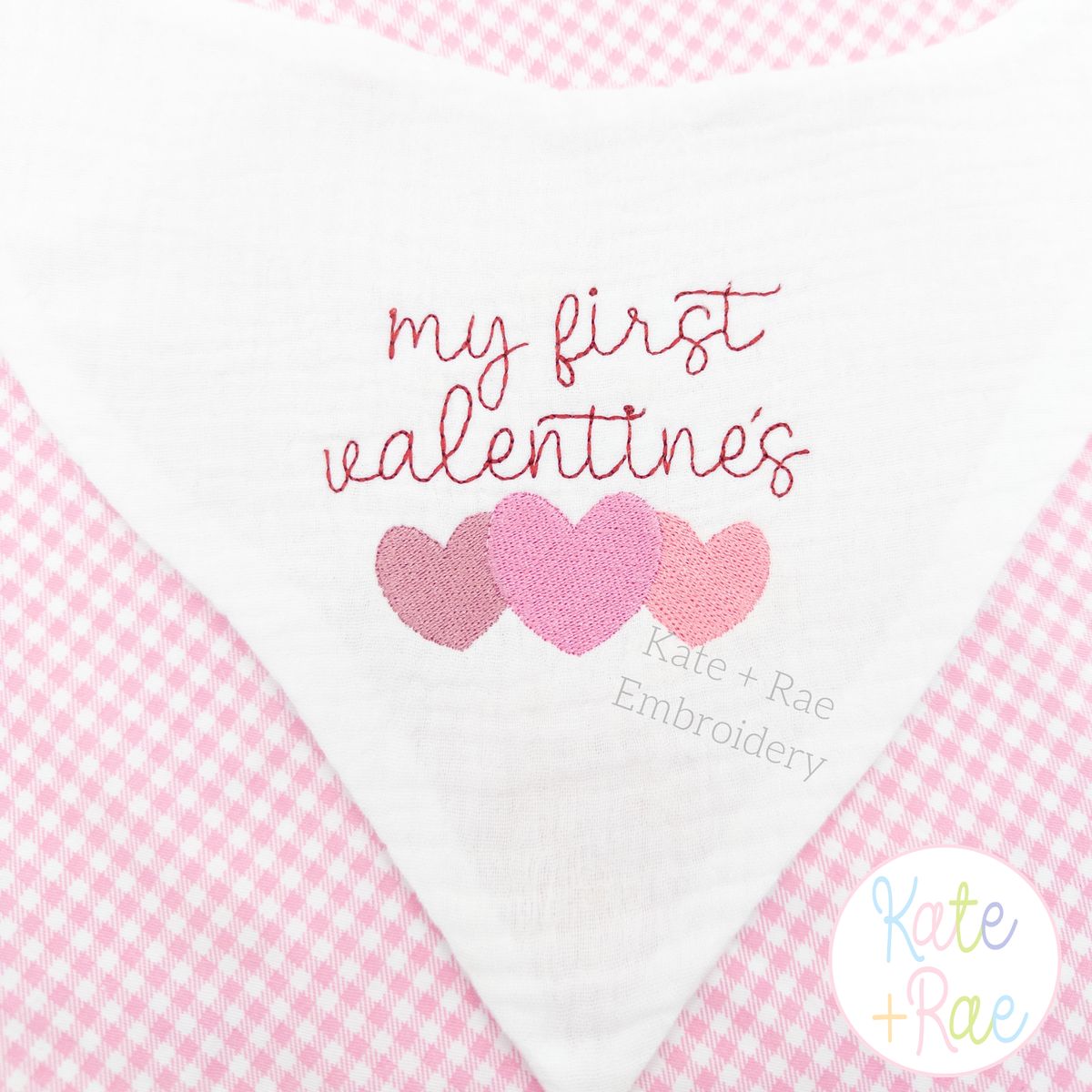 Heart Trio Mini Fill Embroidery Design: Three small pink hearts below the words my first valentine's stitched on a white bib over pink gingham