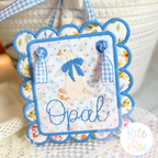 Girl Goose Mini Fill with Bow Embroidery Design: White goose with a blue bow and the name Opal in blue on a scalloped door hanger