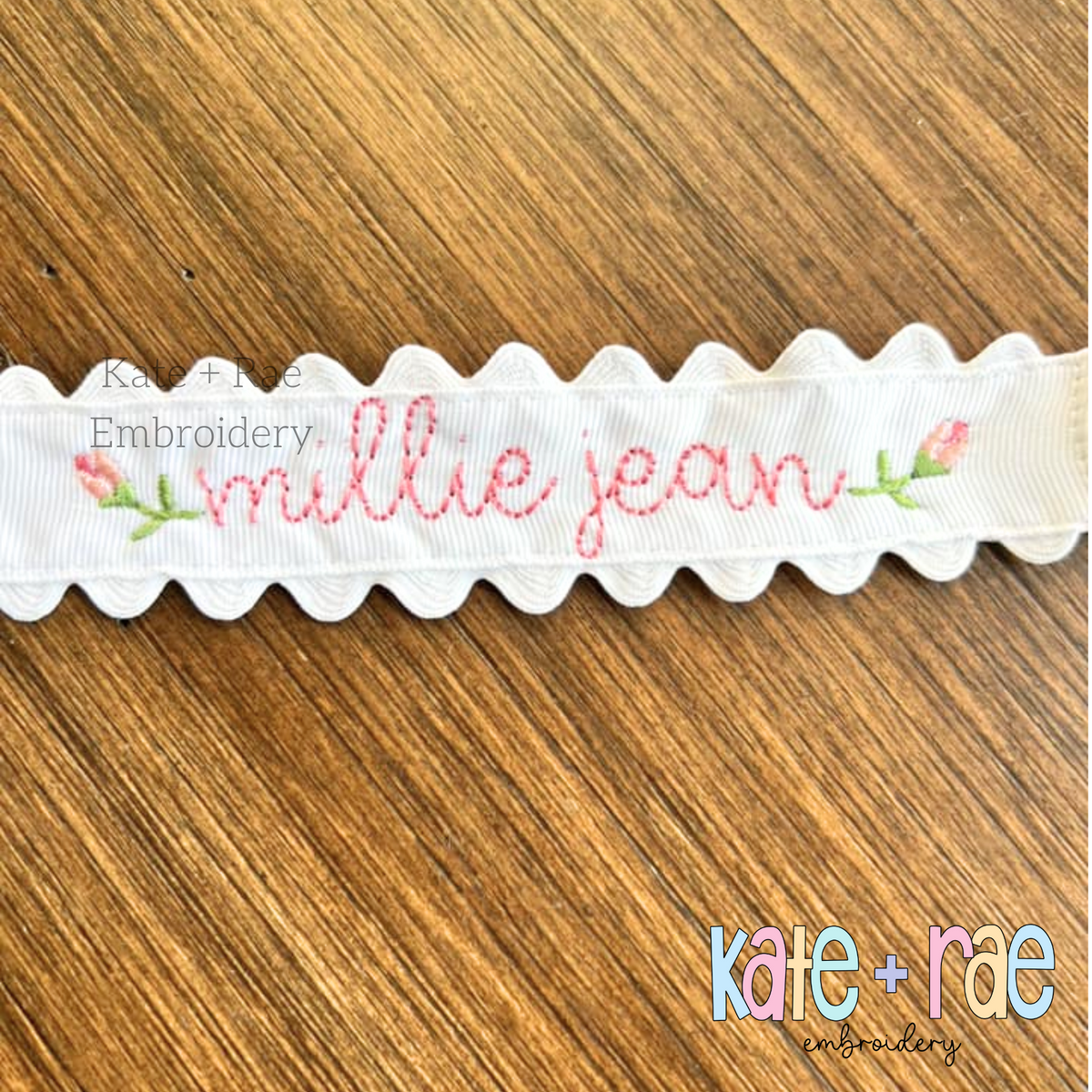 Hello Bean Stitch Embroidery Font: The name millie jean in pink script with rosebuds stitched on a white ric rac headband