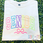 Curved Senior with Bow Satin Stitch Applique Embroidery Design: SENIOR in neon curved collegiate satin applique letters with a pink bow on a cream shirt