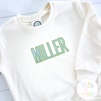 Beau Fill Stitch with Satin Border Embroidery Font: The name MILLER in green and blue fill stitch letters on a cream sweatshirt