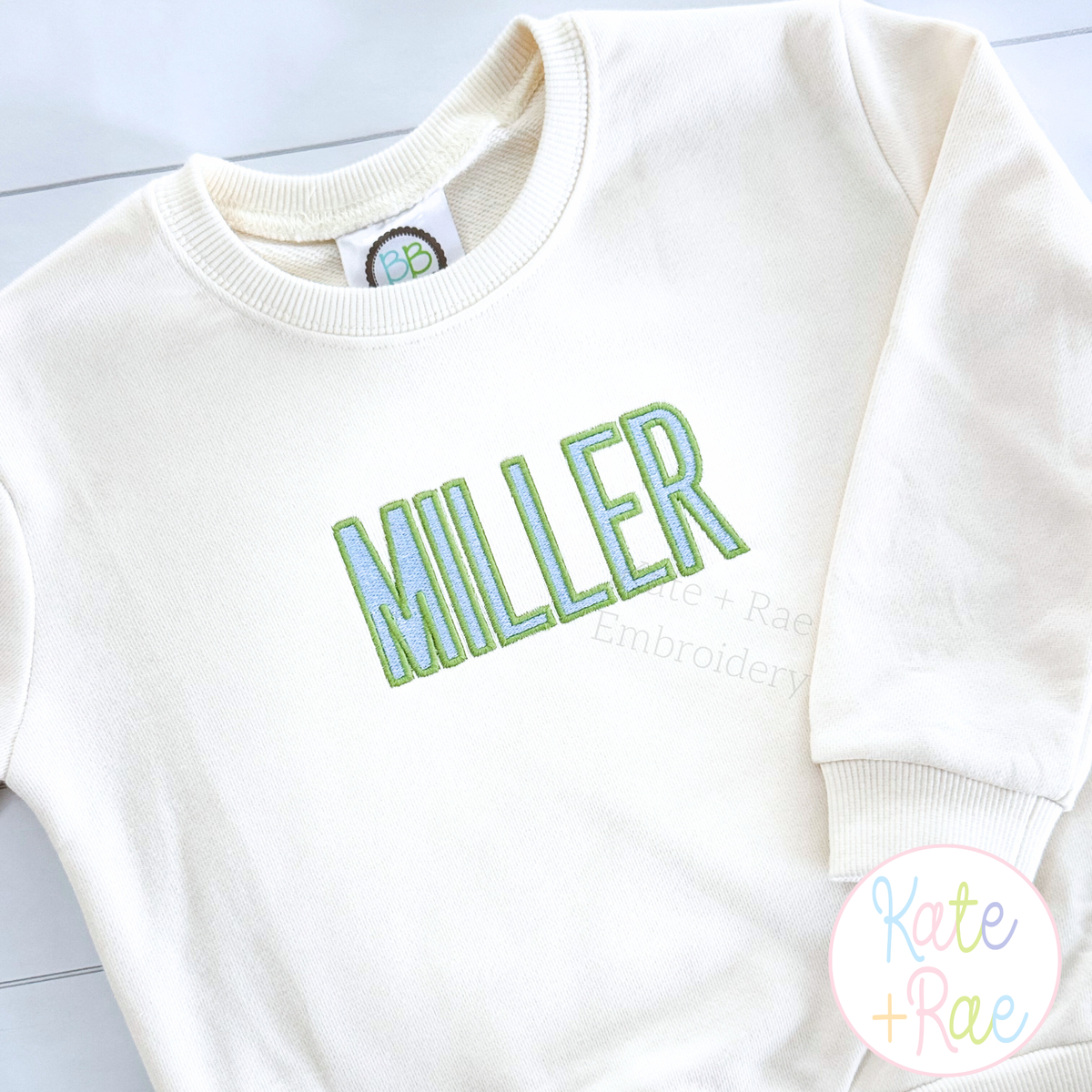 Beau Fill Stitch with Satin Border Embroidery Font: The name MILLER in green and blue fill stitch letters on a cream sweatshirt