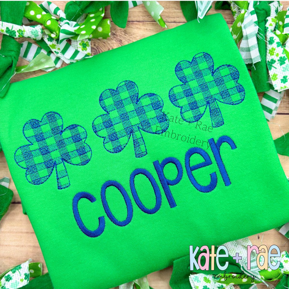 Plaid Shamrock Trio Sketch Stitch Embroidery Design: Three blue gingham sketch stitch shamrocks above the name COOPER in blue on a green shirt