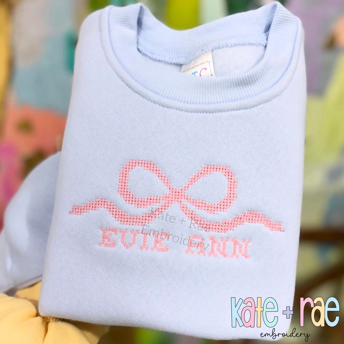 Flowy Bow Cross Stitch Embroidery Design: Pink cross stitch flowy bow above EVIE ANN on a light blue sweatshirt