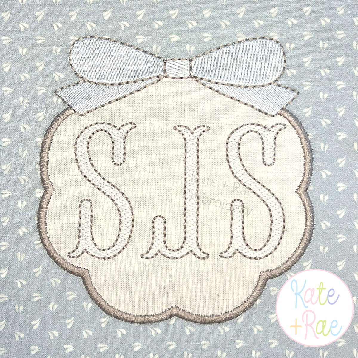 Scallop Circle Monogram Frame with Bow Satin Applique Embroidery Design: Cream scallop frame with a white bow around an SJS monogram on gray print fabric