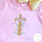 Floral Cross Pattern Fill Stitch Embroidery Design: Tan floral cross with a lavender bow above Matthew 28:6 in lavender script on a pink shirt