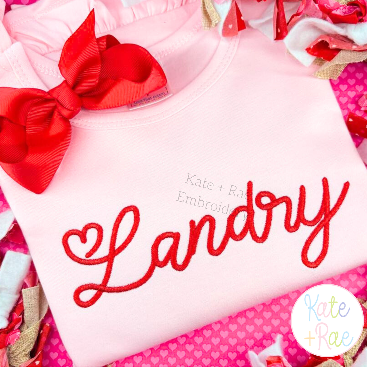 Valentine Heart Satin Stitch Embroidery Font: The name Landry in red heart satin script on a pink shirt