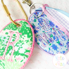 Oval Satin Stitch Bag Tag/ Keychain In The Hoop Embroidery Design: Green and purple floral oval bag tags with pink monograms on gold clips