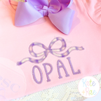 Flowy Longtail Bow Zigzag Stitch Applique Embroidery Design: Purple gingham flowy bow applique above OPAL in lavender on a pink shirt