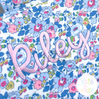Hello Outline Satin Stitch Embroidery Font: The name Riley in pink outline satin script on a blue floral fabric