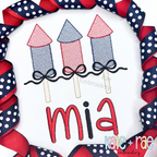 Girl's Firework Trio with Bows Sketch Stitch Embroidery Design: Three red and blue sketch firework rockets with navy bows above the name mia in red on a white shirt