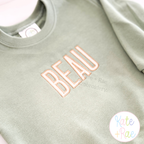 Beau Fill Stitch with Satin Border Embroidery Font: The name BEAU in peach fill stitch letters on a sage sweatshirt