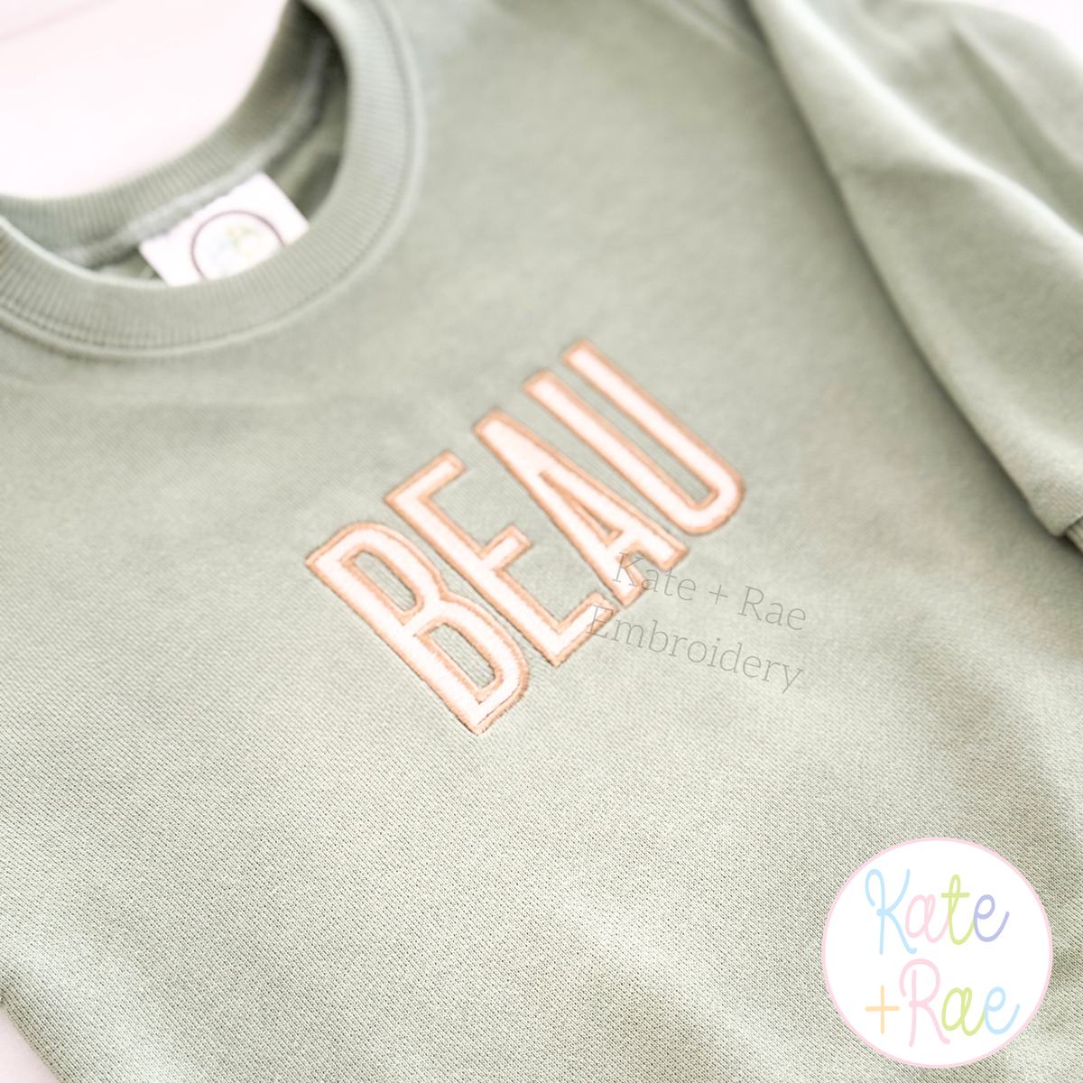 Beau Fill Stitch with Satin Border Embroidery Font: The name BEAU in peach fill stitch letters on a sage sweatshirt