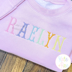 Fishtail Cross Stitch Machine Embroidery Font: The name RAELYN in rainbow fishtail cross stitch letters on a lavender sweatshirt