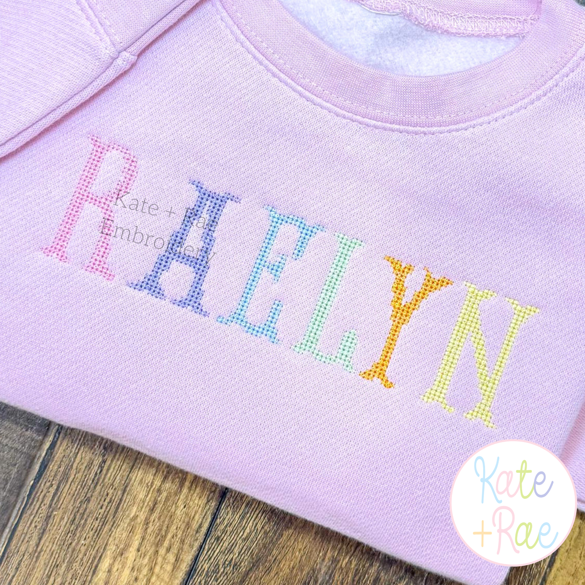 Fishtail Cross Stitch Machine Embroidery Font: The name RAELYN in rainbow fishtail cross stitch letters on a lavender sweatshirt