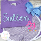 Delilah Two Toned Satin Stitch Embroidery Font: The name Sutton in two toned light blue satin script on a purple shirt