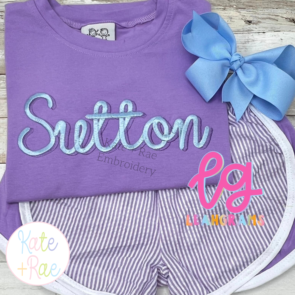 Delilah Two Toned Satin Stitch Embroidery Font: The name Sutton in two toned light blue satin script on a purple shirt