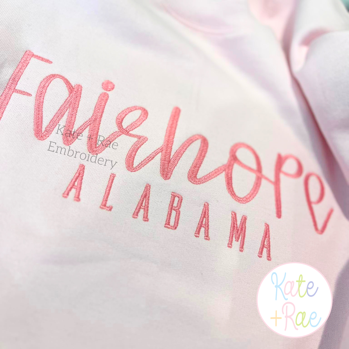Daisy Handwritten Satin Stitch Embroidery Font: Fairhope in pink handwritten script above ALABAMA in pink on a white shirt