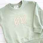 Beau Fill Stitch with Satin Border Embroidery Font: The name BEAU in cream fill stitch letters with tan border on a sage sweatshirt