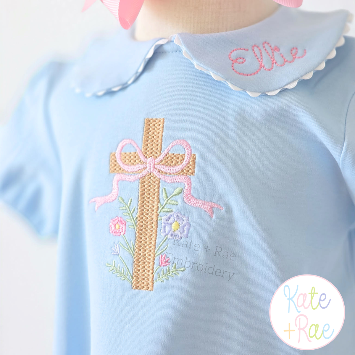 Floral Cross Pattern Fill Stitch Embroidery Design: Tan fill stitch cross with a pink bow and flowers on a light blue shirt with the name Ellie on the collar