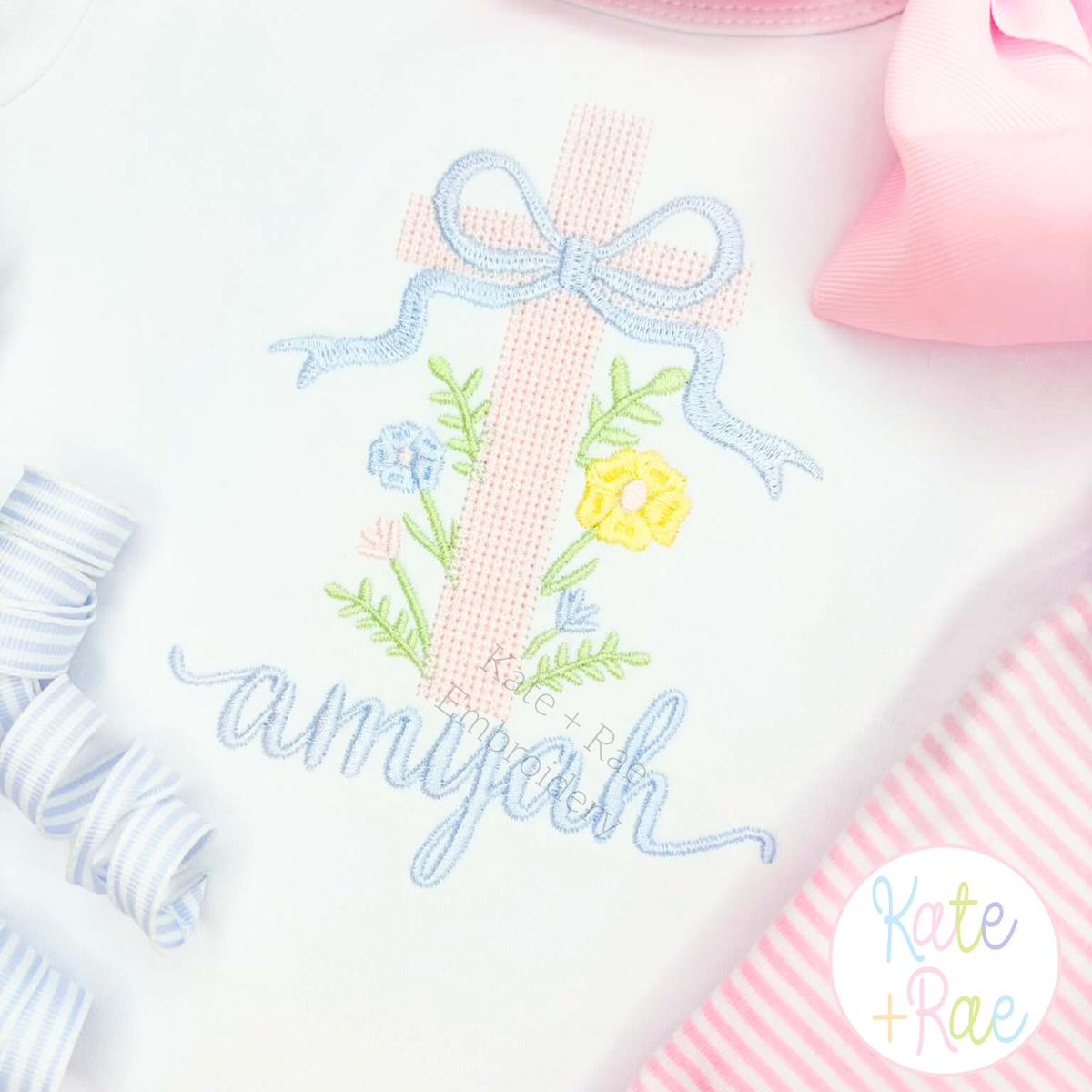 Cross Stitch Floral Cross Embroidery Design: Close-up of a pink cross stitch floral cross with a blue bow above the name amyjah