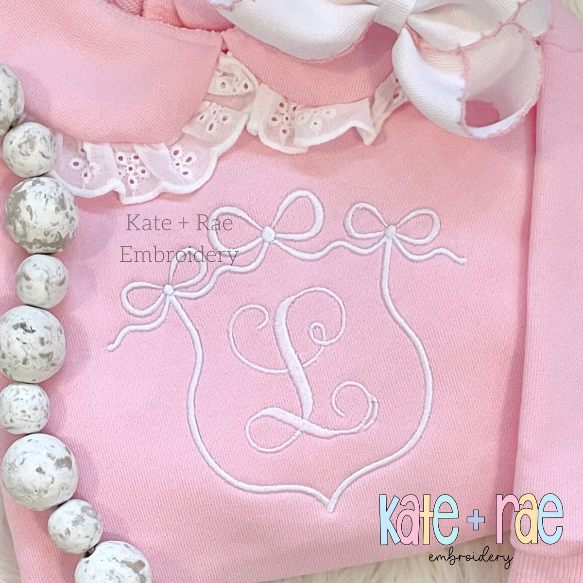 Triple Flowy Bow Crest Embroidery Design: Pink triple bow frame satin stitch outline with a pink script monogram on a lavender sweatshirt