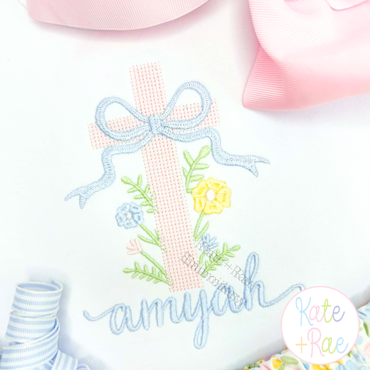Cross Stitch Floral Cross Embroidery Design: Pink cross stitch floral cross with a blue bow above the name amyjah in blue script on white
