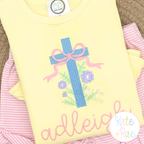 Floral Cross Pattern Fill Stitch Embroidery Design: Blue cross with a pink bow and flowers above the name adleigh in pink script on a yellow shirt