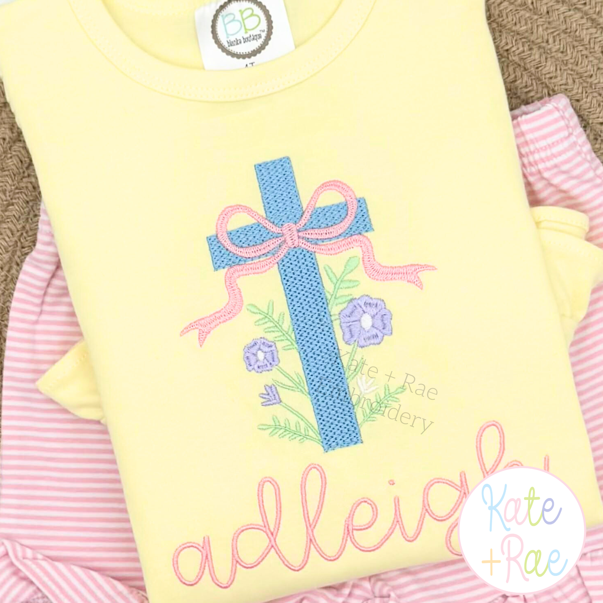 Floral Cross Pattern Fill Stitch Embroidery Design: Blue cross with a pink bow and flowers above the name adleigh in pink script on a yellow shirt