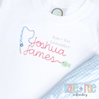 Gone Fishing Bean Stitch Embroidery Font: The name joshua james in pink script with a blue fishing pole and green fish on a white shirt