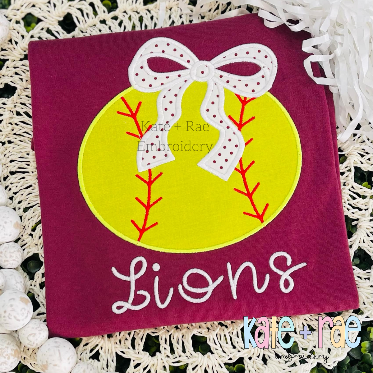 Baseball/ Softball with Merritt Bow Satin Stitch Embroidery Design: Yellow softball applique with a white polka dot bow above the word Lions in white script on a maroon shirt
