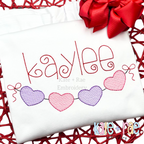 Heart Banner Sketch Stitch Embroidery Design: The name kaylee in red script above a heart banner in purple and pink on a white shirt