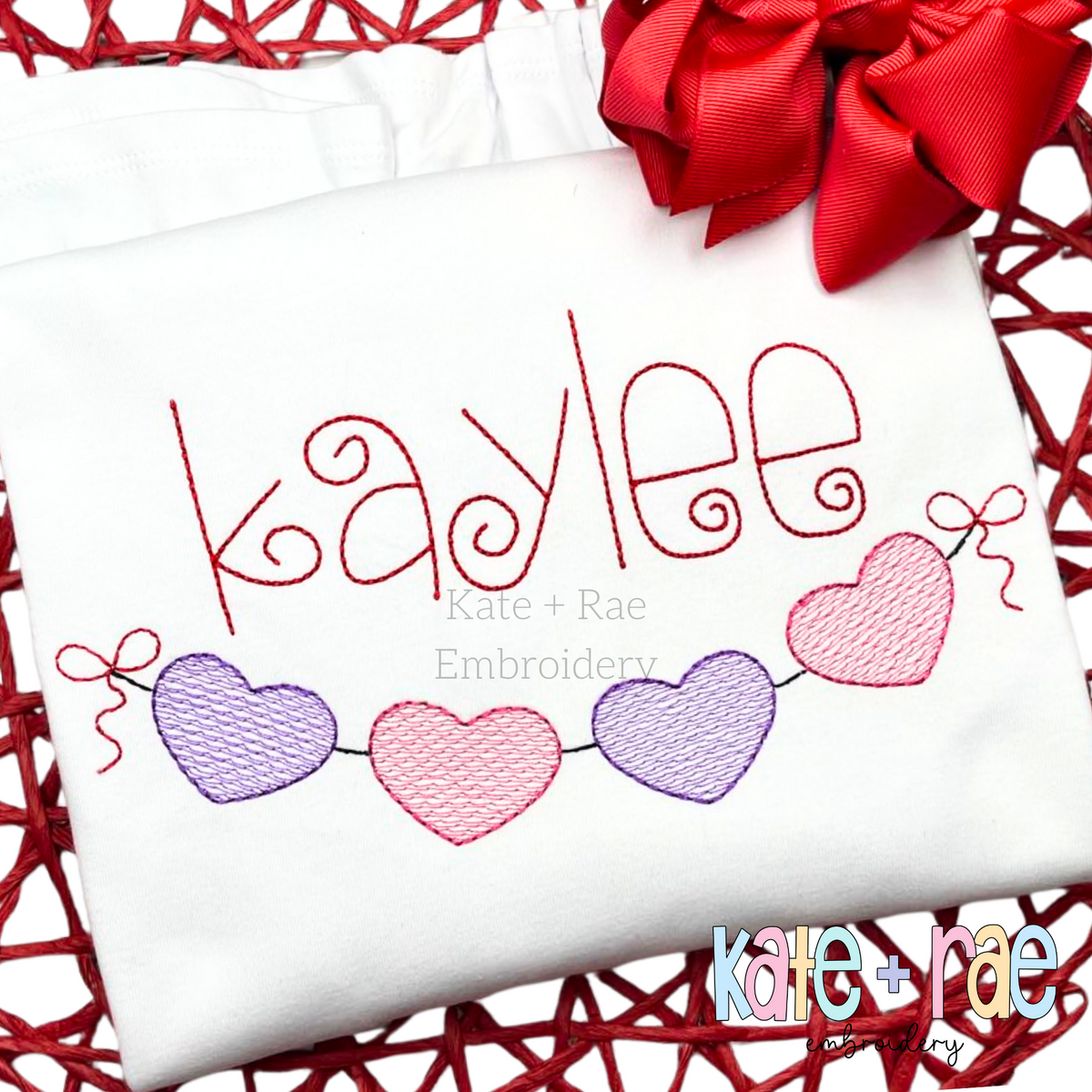 Heart Banner Sketch Stitch Embroidery Design: The name kaylee in red script above a heart banner in purple and pink on a white shirt