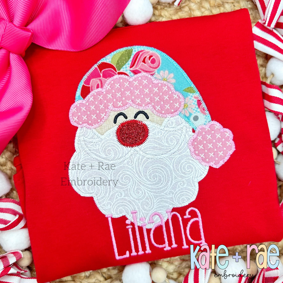 Santa Claus Zigzag Applique Embroidery Design: Santa face applique with a floral hat and red glitter nose above the name Liliana in pink on a red shirt