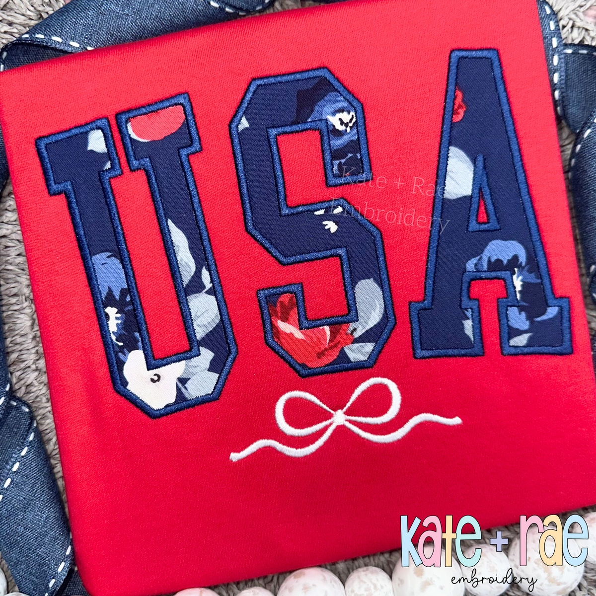 Curved USA with Bow Satin Stitch Applique Embroidery Design: USA in navy floral applique letters with a white bow on a red shirt