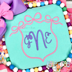 Triple Flowy Bow Crest Embroidery Design: White triple bow frame around a script letter L on a pink sweatshirt
