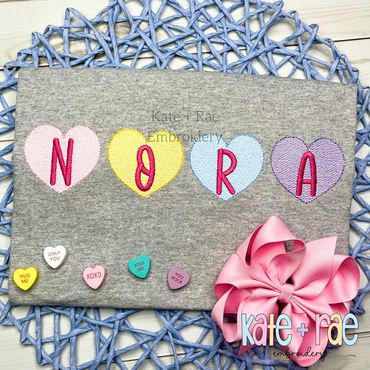 Candy Heart Sketch Stitch Embroidery Font: Pastel candy hearts with pink letters spelling NORA on a gray shirt with mini candy hearts