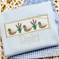 Mallard Ducks and Cattails Sketch Stitch with Satin Applique Patch Embroidery Design: Mallard ducks and cattails on a tan satin applique patch above the name Reid on a light blue shirt