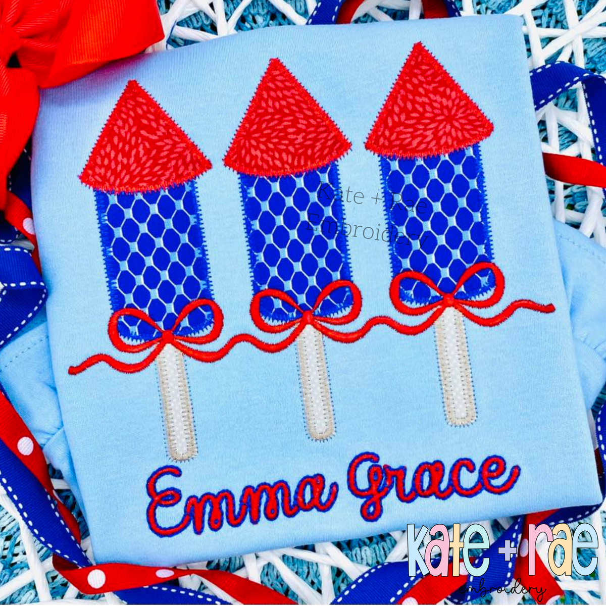 Delilah Outline Satin Stitch Embroidery Font: The name Emma Grace in navy outline script below red and blue firework appliques on a light blue shirt