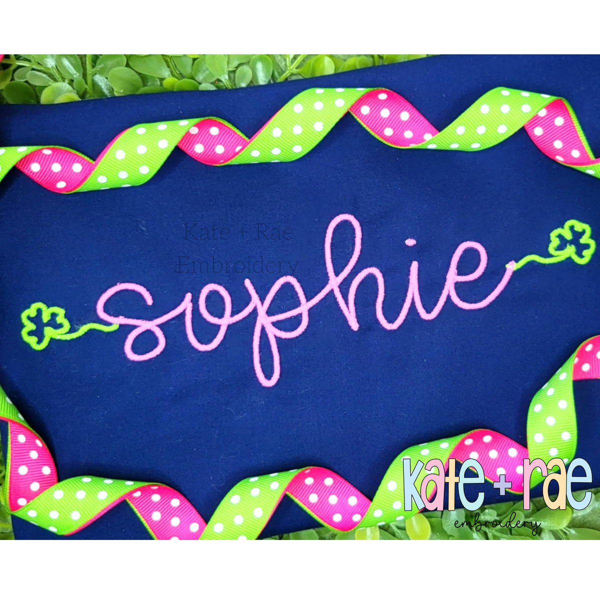 Shamrock Font Add On Embroidery Design: The name sophie in pink script with green shamrock ends on a navy shirt