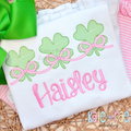 Shamrock Trio with Connecting Bows Sketch Stitch Embroidery Design: Three green sketch stitch shamrocks linked by pink bows above the name Haisley in pink on a white shirt