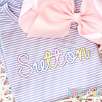 Delilah Outline Satin Stitch Embroidery Font: The name Sutton in pastel outline satin script on a lavender striped shirt