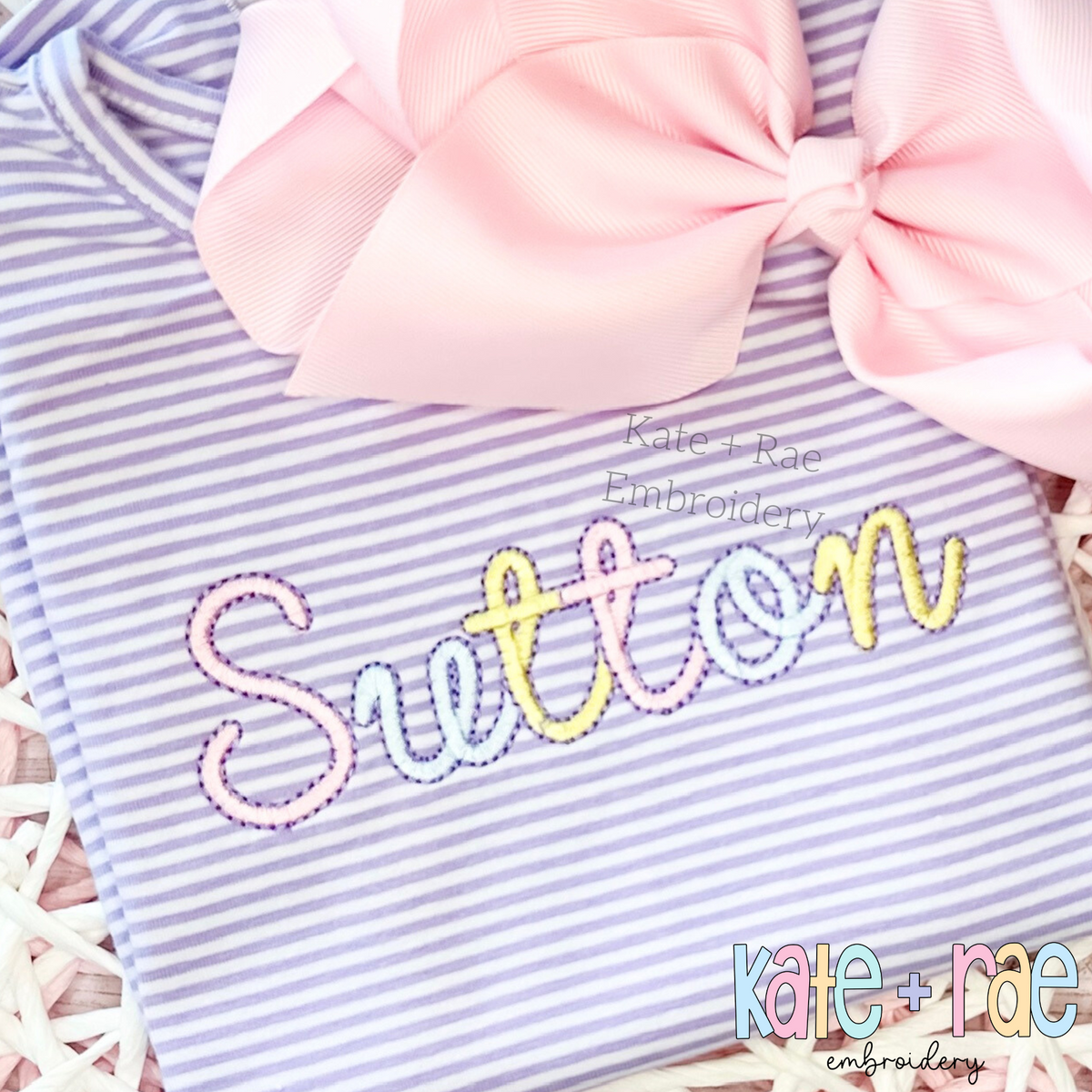 Delilah Outline Satin Stitch Embroidery Font: The name Sutton in pastel outline satin script on a lavender striped shirt