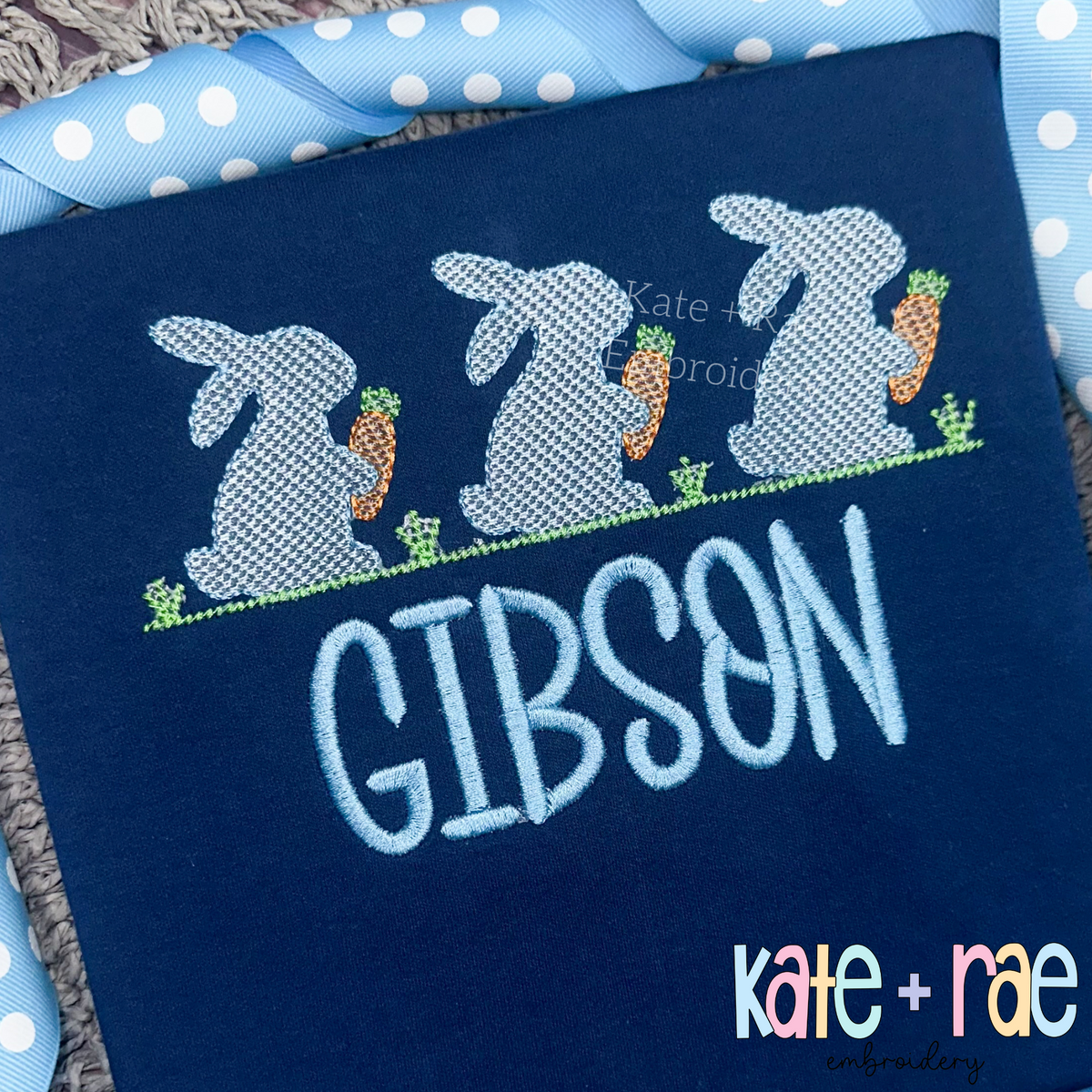 Boy's Easter Bunny with Carrots Trio Sketch Stitch Embroidery Design: Three gray sketch bunnies holding carrots above the name Gibson in light blue on a navy shirt